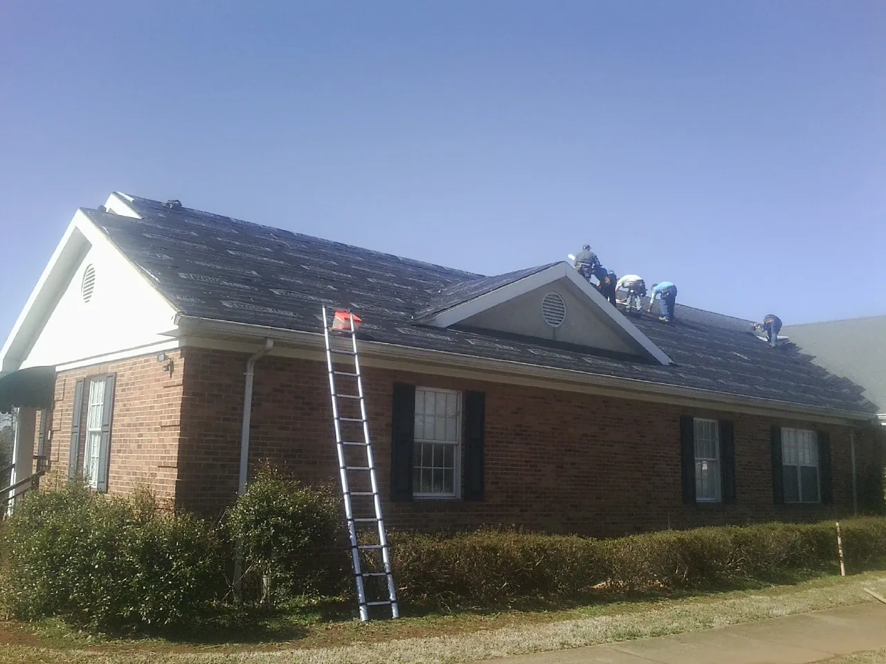 Crew tearing shingles off a brick ranch roof with a ladder set — Emergency Roof Repair work by our crew in Bainbridge