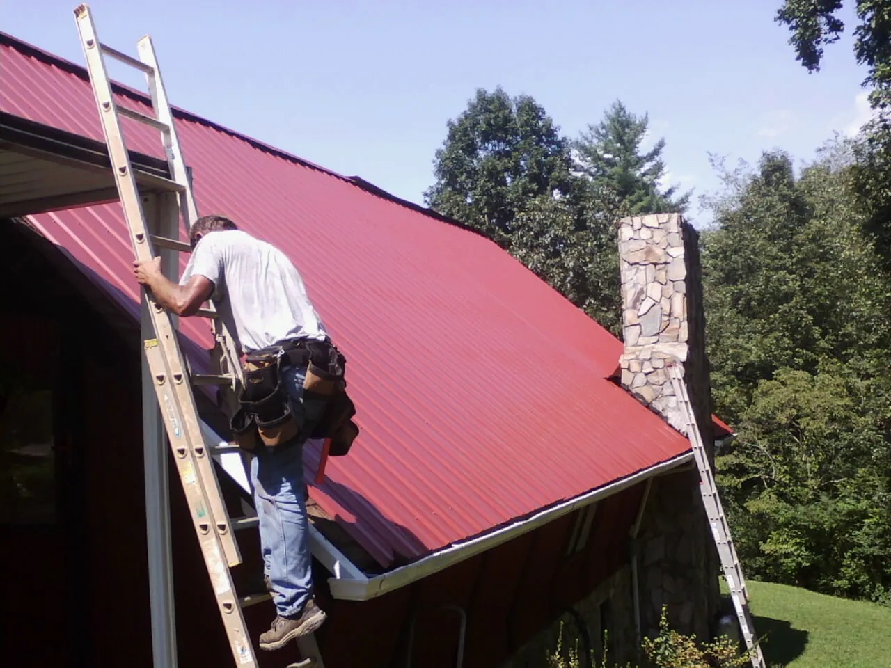 Roofer on a ladder beside a red metal roof and stone chimney — Soffit & Fascia Repair by Roofing Brothers in Bainbridge